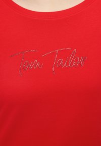 Κόκκινο ύφασμα με το κείμενο „Tom Tailor“ γραμμένο με μικρές λαμπερές στρας στην περιοχή του στήθους.