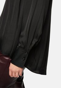 Schwarze, durchsichtige Bluse mit langen Ärmeln, ausgestattet mit einem Knopfsaum, der mit quadratischen schwarzen Akzenten verziert ist. Glatte Textur mit einem fließenden Design.