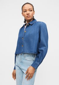 Νέα γυναίκα με πλεξούδες, που φοράει ένα κοντό τζιν μπουφάν με κουμπιά και ανοιχτόχρωμο τζιν ψηλόμεσο παντελόνι, κοιτάζοντας προς τα πλάγια.