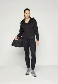 Sort zip-up hoodie med hætte, parret med sorte leggings og grå sportssko. Model holder en sort duffelbag.