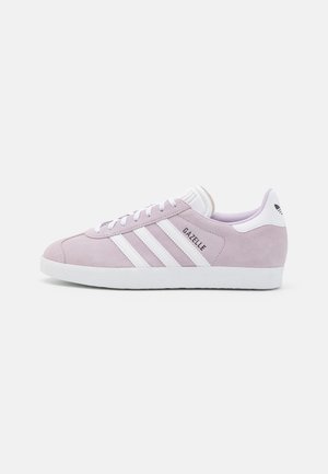 Jasnofioletowy sneaker Adidas Gazelle z białymi paskami, białą podeszwą i białą zakładką na pięcie z czarnym logo Adidas, widoczny z profilu.