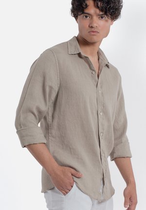 Giovane uomo che indossa una camicia beige con bottoni e maniche risvoltate fino ai gomiti e pantaloni bianchi, in piedi con una mano in tasca su sfondo bianco.