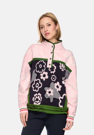 Frau trägt einen rosa-schwarzen Pullover mit Blumenmuster, grünem Besatz und geknöpftem Kragen, steht vor einfarbigem Hintergrund.