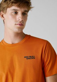 Orange bomulds t-shirt med et sort "RED BULL RACING" logo på brystet, rund halsudskæring og korte ærmer. Blød tekstur.