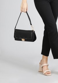 Borsa a mano in pelle nera con dettaglio arricciato sul davanti, chiusura in oro e tracolla removibile, tenuta da una persona in pantaloni neri.