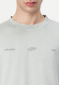 Lichtgrijze t-shirt met een ronde hals, voorzien van subtiele branding in grijze opdruk op de voorkant. De stof heeft een gladde, zachte textuur.