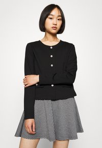 Fekete kardigán kerek nyakkivágással, elülső gombos zárral és két zsebbel, fekete-fehér kockás keresztbe szabott szoknyával párosítva.