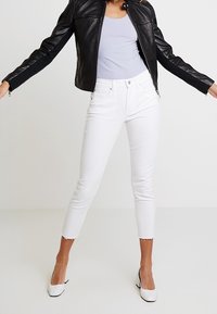 Vaqueros blancos a la altura del tobillo con dobladillo desflecado, combinados con una camiseta sin mangas gris claro y una chaqueta de cuero negra, usados con zapatos de tacón altos blancos.