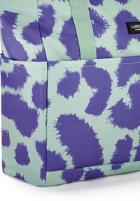 Zöldeskék táska közelről, purple szabálytalan foltokkal, elülső zsebbel és zöldeskék fogantyúval.