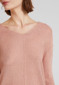 Jasnoróżowy sweter z dzianiny z dekoltem w kształcie V, charakteryzujący się luźnym fasonem i teksturowanym materiałem. Szew biegnie pionowo wzdłuż środka.