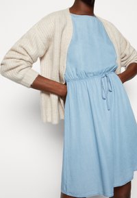 Světle modré denimové šaty s elastickým pasem a stažením pomocí šňůrky; doplněné krémovým pleteným kardiganem s žebrovanou texturou a otevřeným předním dílem.