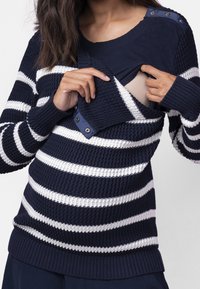 Žena v námořnickém modrém a bílém pruhovaném svetru s bočním zapínáním pro kojení, který je odklopený, aby odhalil béžovou podprsenku.