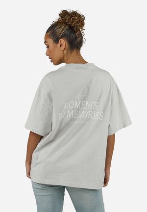 Göndör hajú nő kontyba tűzve, világosszürke, túlméretezett pólót visel, amelynek hátán a "Moments to Memories" felirat olvasható, valamint világoskék farmert.