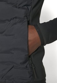 Sort polstret jakke med en glat overflade, lynlåslomme og tætsiddende ærmer i strækstof for komfort og bevægelsesfrihed.