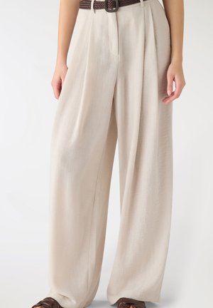 Pantaloni beige a vita alta con gamba larga e pieghe frontali, abbinati a una cintura intrecciata marrone e scarpe slip-on in pelle marrone.