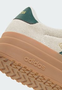 Sneakers Adidas con una toma in suede color crema, dettagli verdi, una suola texturizzata color tan e branding in rilievo. Design curvo con motivo di battistrada visibile.