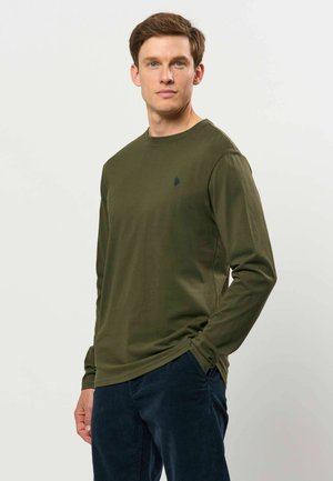 Jeune homme portant une chemise à manches longues vert olive et un pantalon bleu marine, debout avec une main dans la poche devant un fond uni.