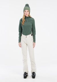 Giacca in pile verde con zip, pantaloni beige con bottoni e scarponi da sci neri. Il modello indossa un berretto lavorato a maglia verde con dettaglio pom-pom.