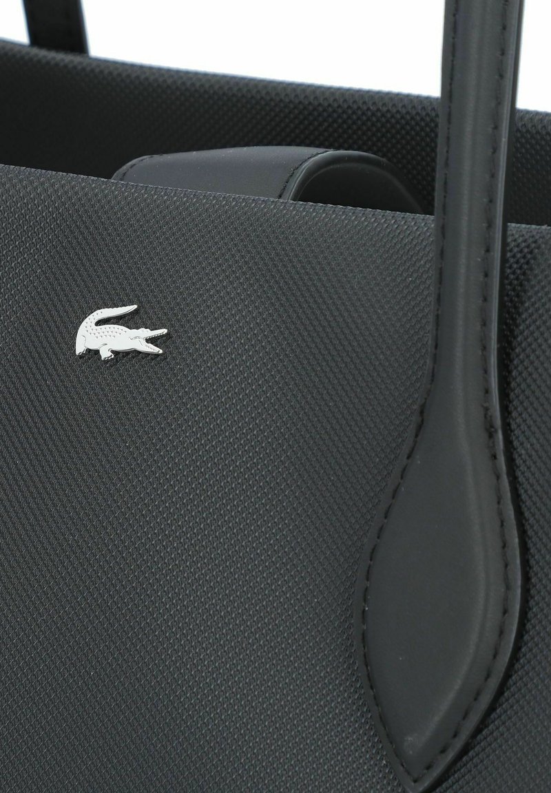 Sac fourre-tout noir texturé avec une anse en cuir lisse et un accent logo crocodile argenté sur le devant.