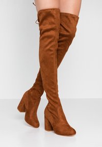 Femme portant des cuissardes en daim brun à talons blocs, jambes croisées, sur un fond clair uni.