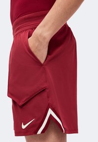 Personne portant un short de sport bordeaux avec une main dans la poche latérale et un logo Nike blanc près de l'ourlet.