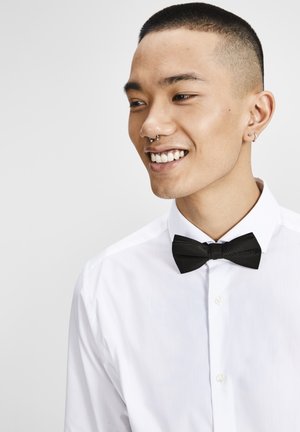 Camisa blanca de botón con una pajarita negra texturizada. La camisa tiene un collar francés y no presenta patrones visibles. Textura de tela suave.