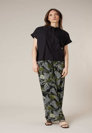 Femme portant une chemise noire à manches courtes boutonnée et un pantalon à jambes larges avec des motifs de feuilles vertes et de léopard blanc, posant devant un fond uni.