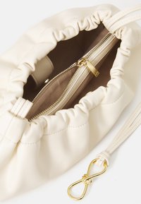 Cremefarvet lædertaske med en samlet top, der viser en lynlåslomme og gyldne hardware-detaljer. Indeni har den en beige foring.