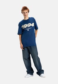 Ung man med ljust brunt hår som bär en blå oversized sporttröja från 1994, stora mörka jeans och vita sneakers, stående avslappnat.