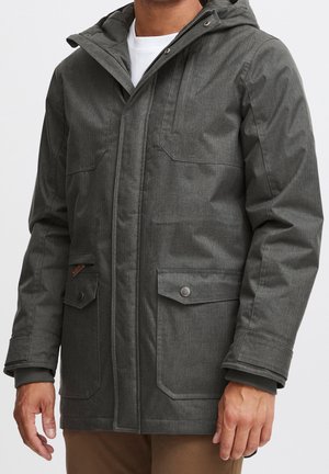 Mann trägt eine dunkelgraue Kapuzenjacke mit aufgesetzten Taschen mit Knöpfen über einem weißen Hemd und brauner Hose.