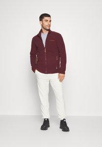 Giacca in fleece bordeaux con tasche con zip, colletto alto e dettagli arancioni, abbinata a pantaloni crema e scarpe da trekking nere.
