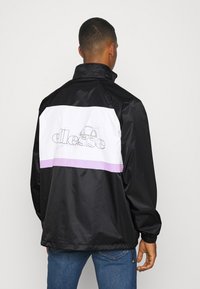 Schwarze Jacke mit weißen und lavendelfarbenen Streifen, ausgestattet mit einem hohen Kragen und elastischen Bündchen. Logo deutlich auf dem Rücken platziert.