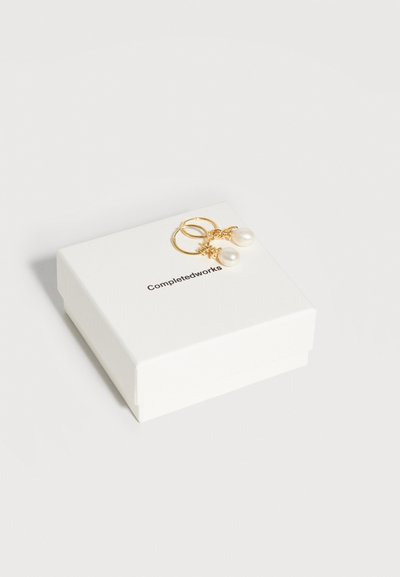 Χρυσά σκουλαρίκια κρίκοι με κρεμαστά λευκά μαργαριτάρια πάνω σε λευκό κουτί με την ετικέτα «Completedworks» σε ανοιχτό γκρι φόντο.