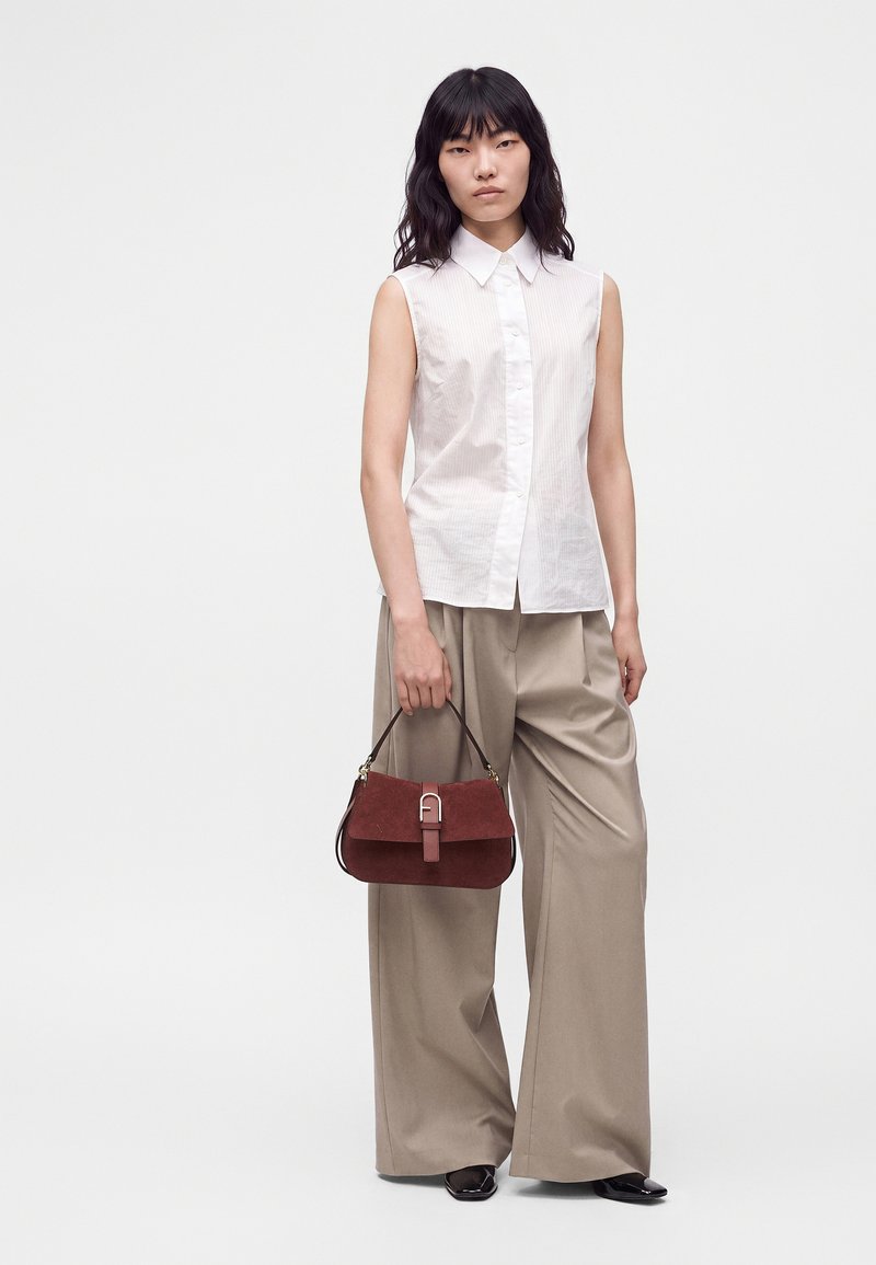 Femme aux cheveux noirs portant une chemise blanche sans manches boutonnée et un pantalon large beige, tenant un petit sac à main marron dans sa main droite.