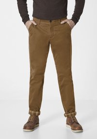 Braune Baumwollchinos mit geradem Bein, ausgestattet mit vorderen Taschen, einem Knopfverschluss und karierten Saumabschlüssen. Getragen mit braunen Lederstiefeln.