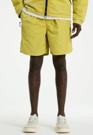 Férfi sárga rövidnadrágban és hozzáillő dzsekiben, fehér sportcipőkkel, fehér háttér előtt állva.