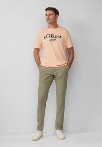 Perzikkleurige t-shirt met zwart logo, lichtgroene broek en witte sneakers. T-shirt heeft een ronde hals; de broek heeft zakken en een gladde textuur.