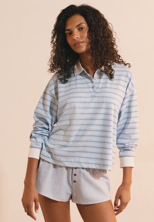 Giovane donna con capelli ricci indossa una camicia a righe azzurro chiaro a maniche lunghe e pantaloncini chiari con bottoni, in piedi davanti a uno sfondo semplice.