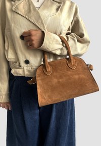 Borsa in suedè marrone con manici arrotondati e forma strutturata, tenuta in mano, abbinata a un giubbotto beige chiaro e pantaloni blu scuro.