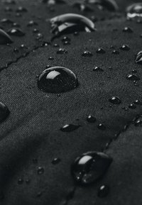 Black waterproof fabric with water droplets on the surface, highlighting texture and sheen. Stitches visible in the background.