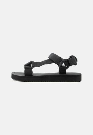 Black flat sandal with adjustable straps over the toes, ankle, and heel, featuring a textured sole and buckle closures.
