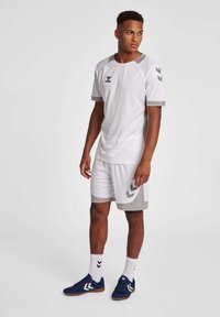 Hvid atletisk skjorte med grå detaljer, parret med hvide shorts med grå kant. Modellen har navy sko og hvide sokker med sorte accenter på.