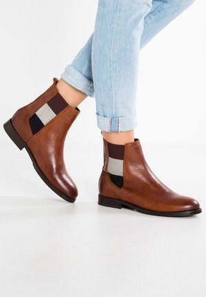 Bottines en cuir marron avec panneaux élastiques sur les côtés en noir, blanc et bordeaux, portées avec un jean bleu clair retroussé sur fond blanc.