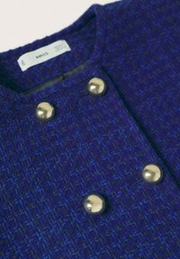 Veston bleu en tissu tissé avec un motif texturé, boutons métalliques ronds et encolure arrondie. Étiquette visible au col.