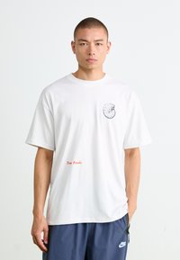 Young man with short hair wearing a white T-shirt with a fossil graphic and red text "Texas Fossils," paired with blue Nike pants.