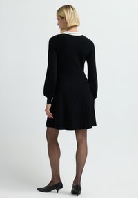 Vestito nero a maglia con maniche lunghe, colletto bianco e gonna plissè. Indossato con scarpe nere e collant trasparenti.