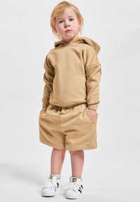 Sudadera beige con capucha y hombros caídos, combinada con pantalones cortos a juego. Zapatillas blancas con rayas negras completan el atuendo.
