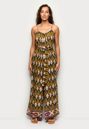 Žena stojící v bezrukávém vzorovaném overal s vázaným páskem, širokými nohavicemi a sandály na jednobarevném světlém pozadí.