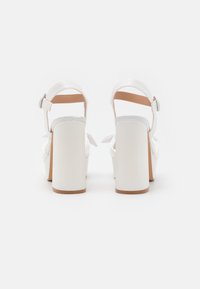 Sandalias blancas de tacón alto con tacón cuadrado y correa en el tobillo. Material superior liso y interior en tono bronceado, con un detalle de lazo en la parte posterior.