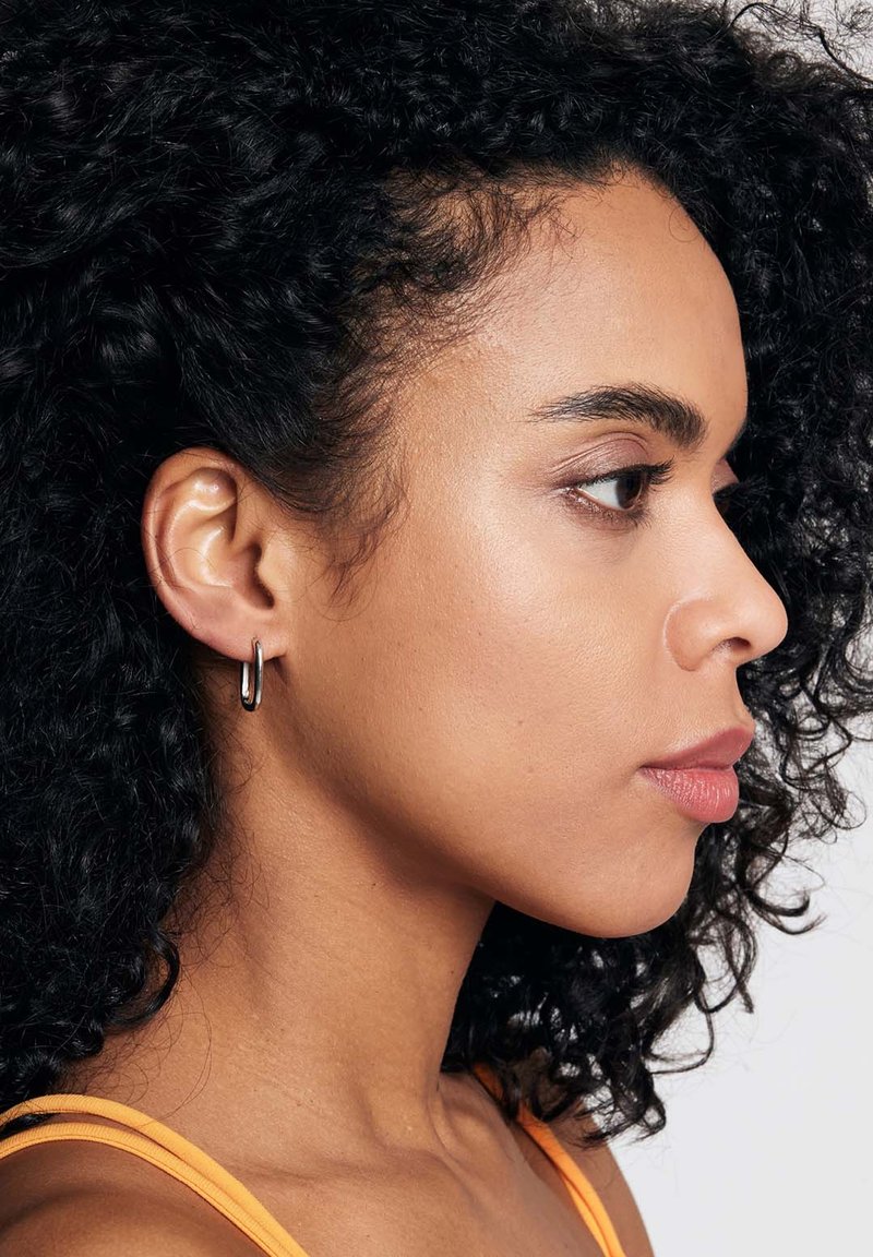 Srebrna kolczyk na kółku przedstawiona na modelce z kręconymi włosami. Kolczyk jest smukły, cylindryczny o polerowanej powierzchni, co podkreśla ogólny design.